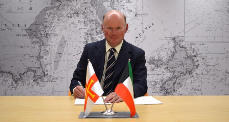 Director General William Mason signing the MoU at a desk behind Guernsey and Kuwait flags